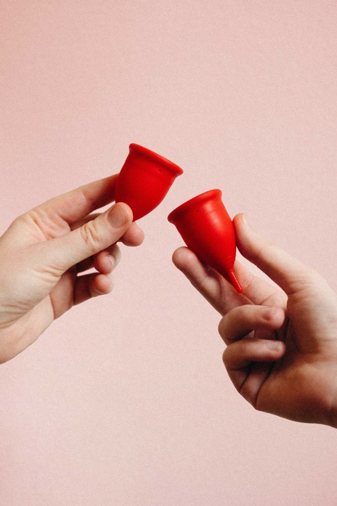 Red period cups with hands holding them in front of a pink background.